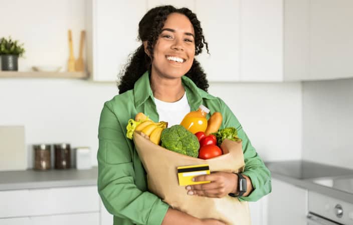 Mulher sorridente segurando sacola com frutas e verduras e um cartão nas mãos, representando o benefício do vale-alimentação para empregadas domésticas.