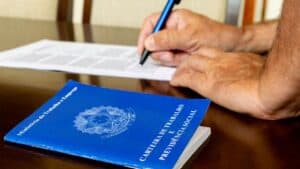 Carteira de Trabalho e Previdência Social sobre a mesa ao lado de uma pessoa assinando um contrato, representando o processo de registro formal de um empregado doméstico.