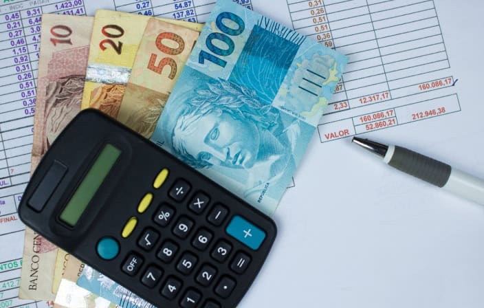 Cédulas de real ao lado de uma calculadora, caneta e planilhas financeiras, representando o cálculo e recolhimento do INSS das empregadas domésticas.